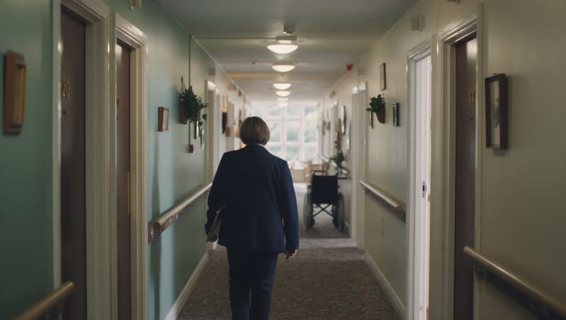 A woman carrying a clipboard walking away down a long care home corridor lined with handrails and bedroom doors.