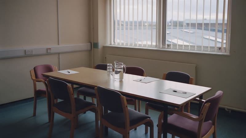 An empty meeting room in a hospital administration block, with notepads, pens and a water jug set out on a long table and vertical blinds half open onto a flat roof.